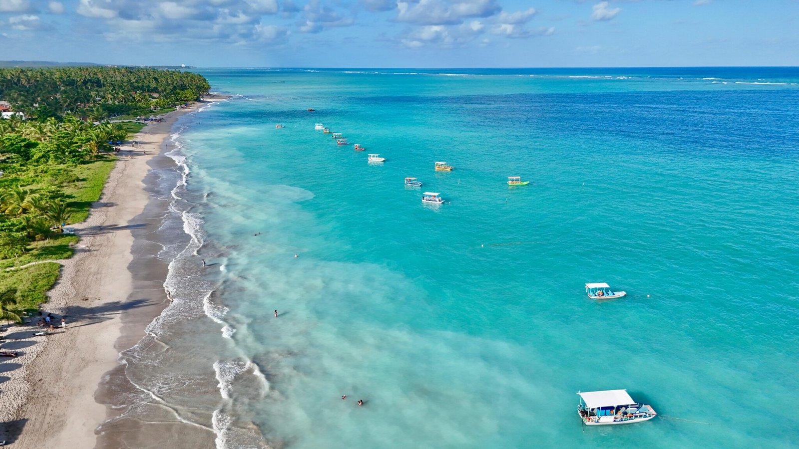 Vista aérea de drone da praia de Maragogi, Alagoas, com mar azul-turquesa, coqueiros e embarcações nas águas cristalinas.